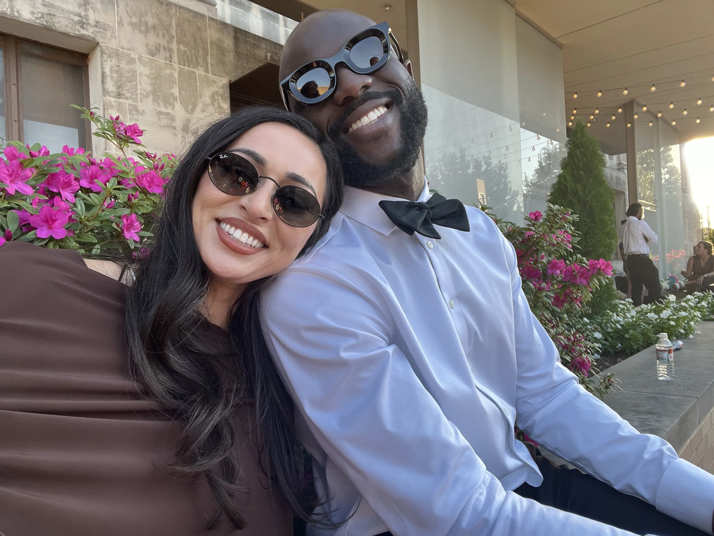 Smiling couple wearing sunglasses pose for a selfie beside vibrant pink flowers at an outdoor patio.
