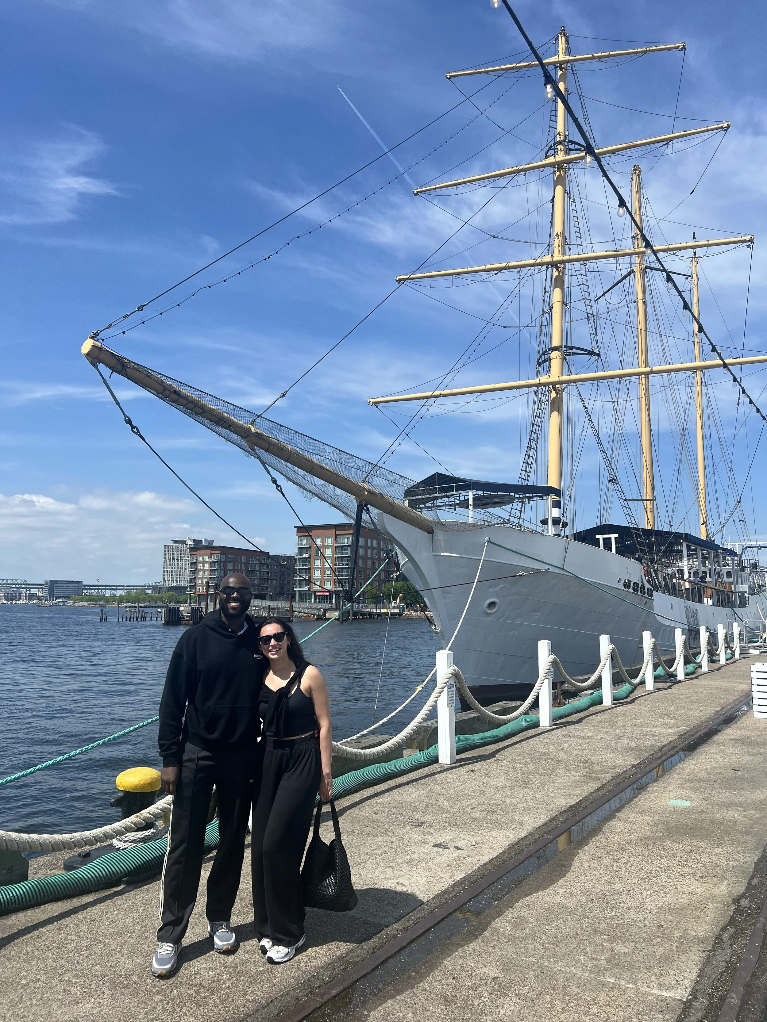 Two people pose beside a large historic sailing ship at a waterfront dock under a bright blue sky.