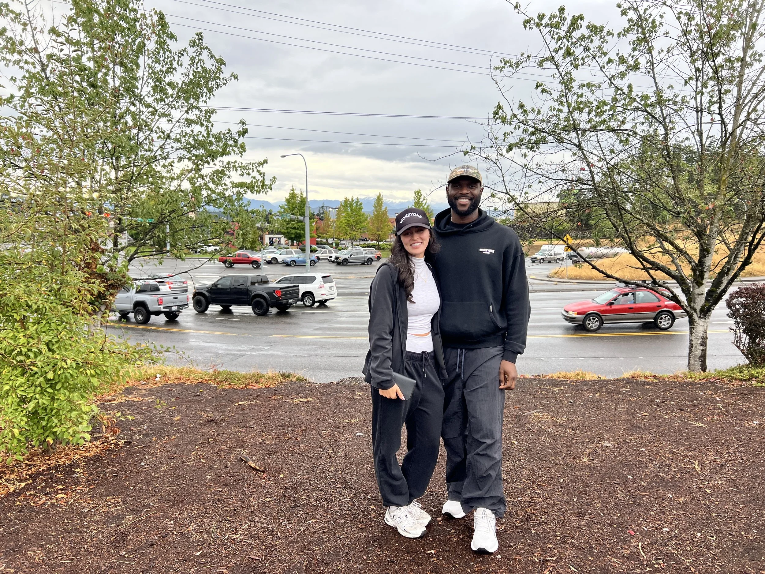 Smiling couple wearing matching black outfits and caps pose in a landscaped area beside a busy road, with trees and mountains in the background.
