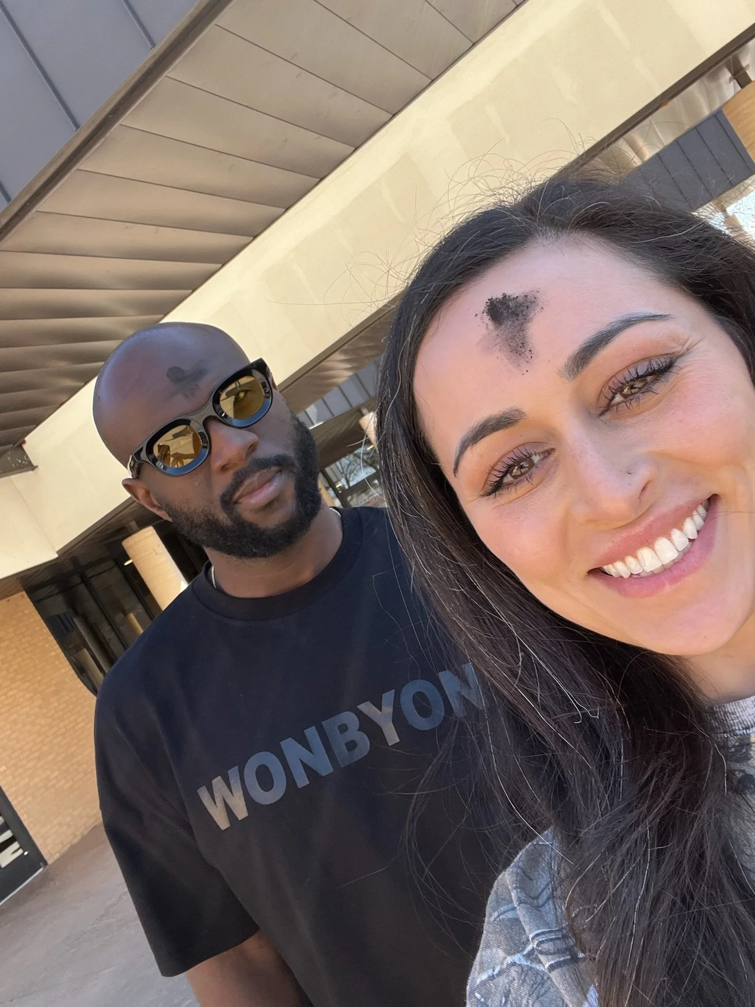 Close-up selfie of two smiling people outdoors, with a woman in the foreground and a man wearing sunglasses behind her.