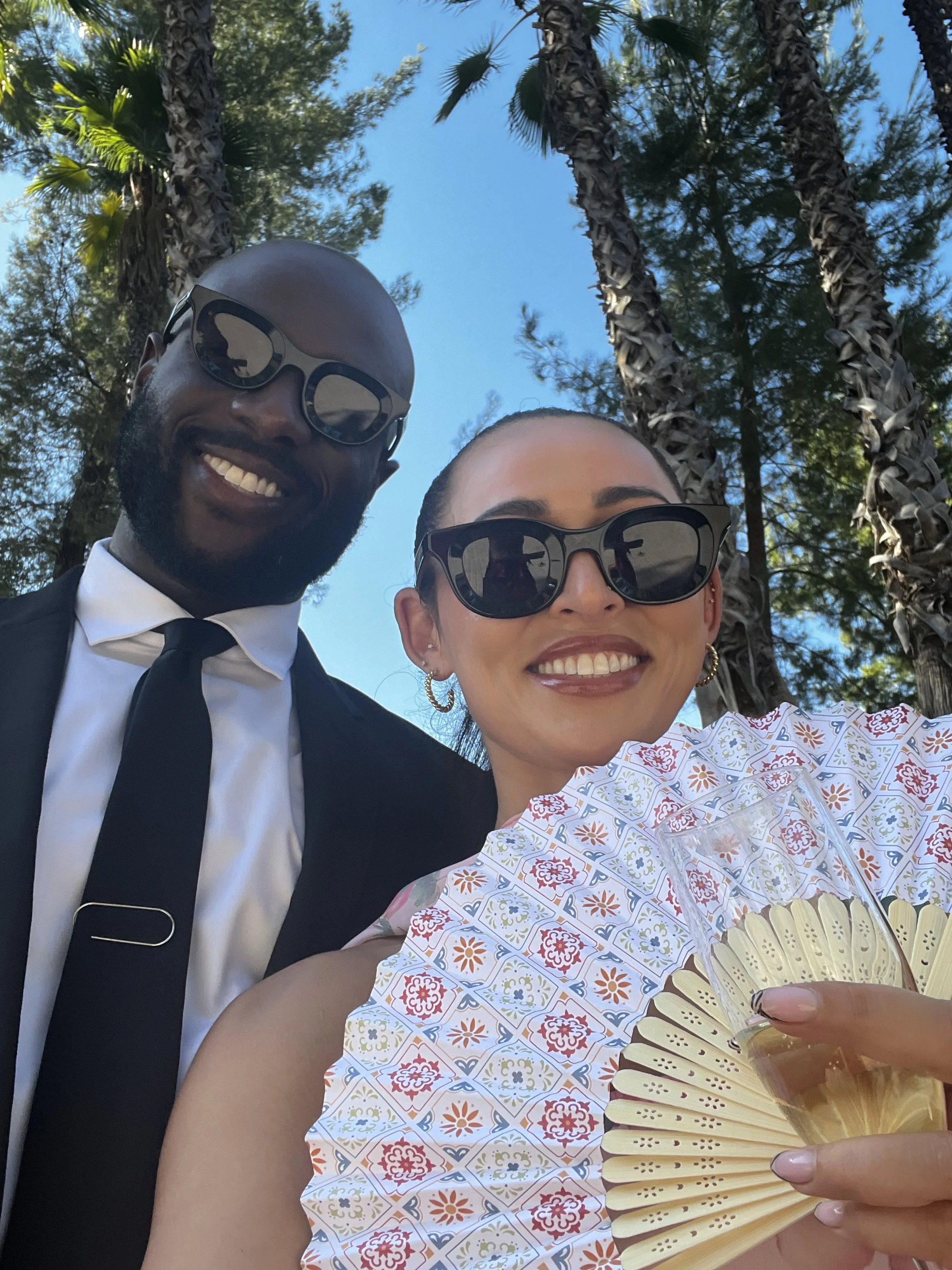 Smiling couple wearing sunglasses outdoors among palm trees, with the woman holding a folding fan.
