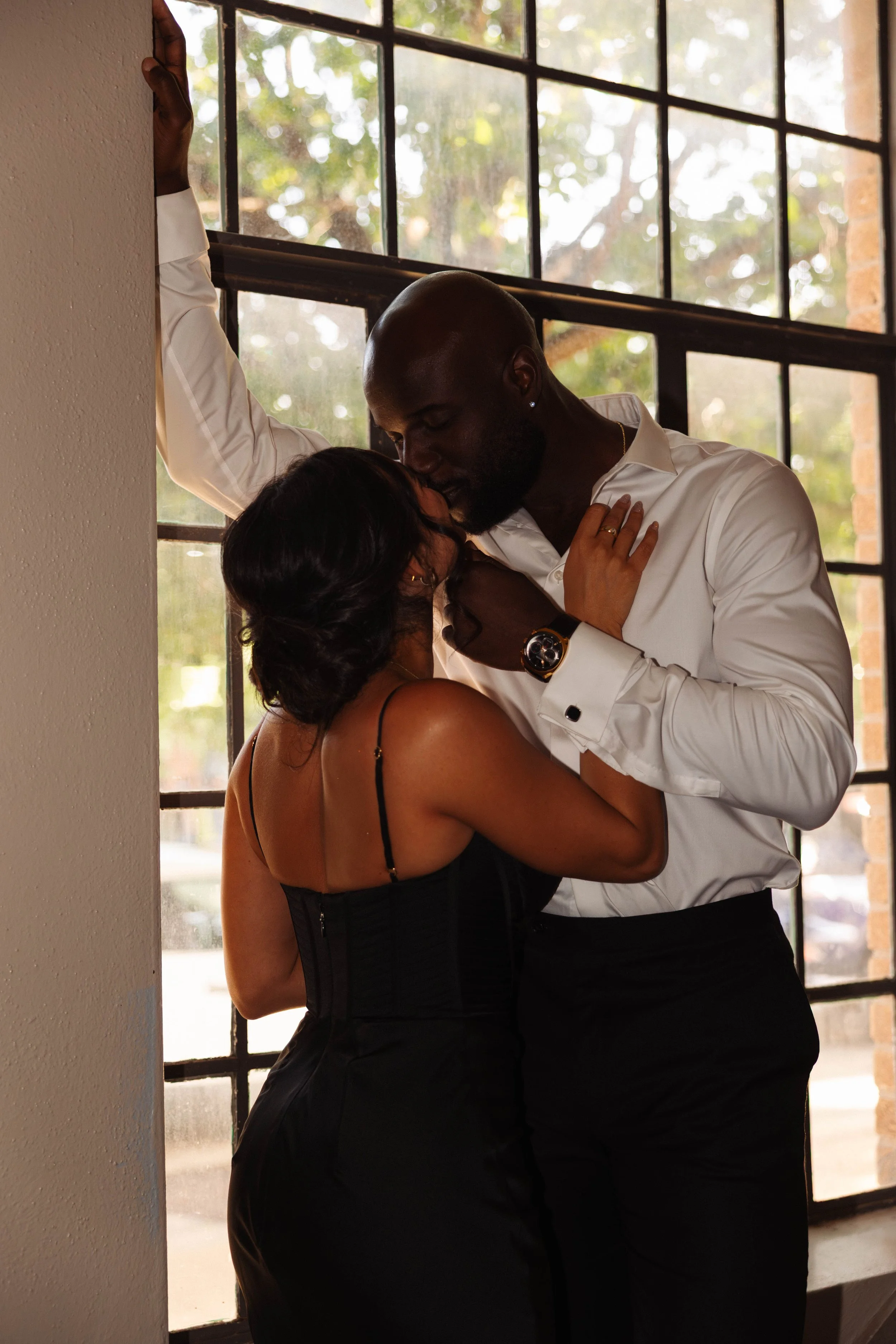 Couple embracing by a sunlit window, with the man in a white shirt and the woman in a black dress.