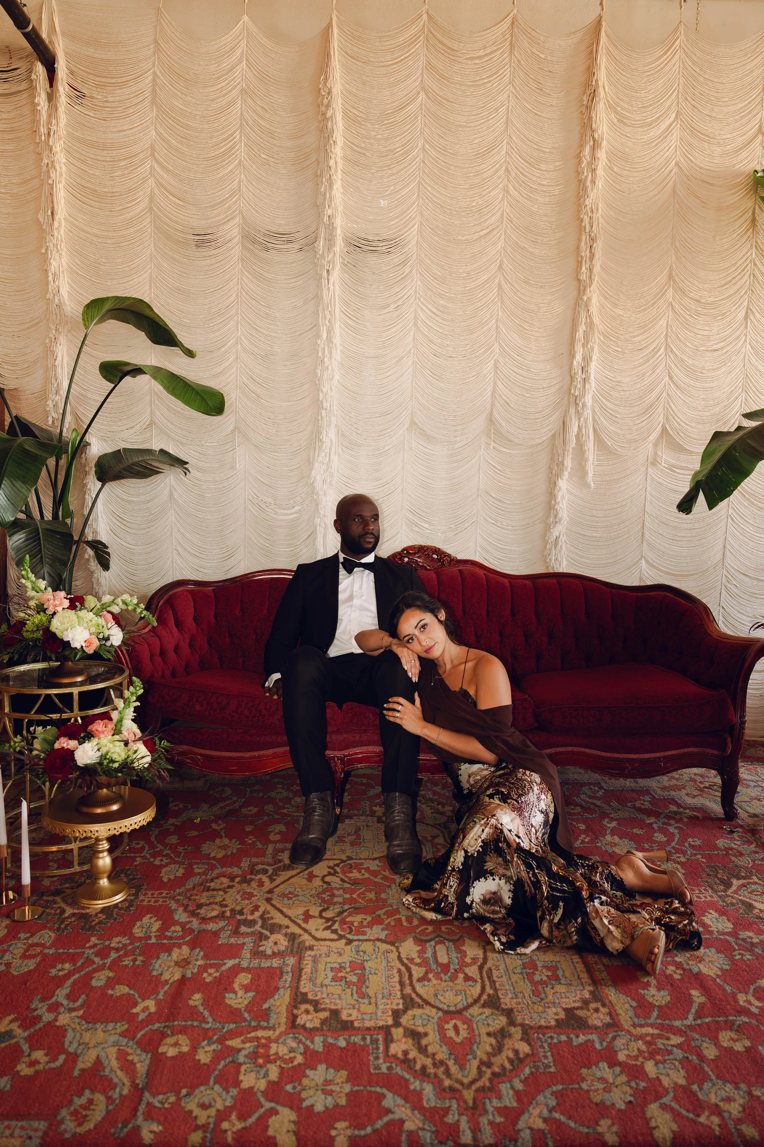Well-dressed couple posing on a red velvet sofa in an elegant room with draped fabric, tropical plants, flowers, and a patterned carpet.