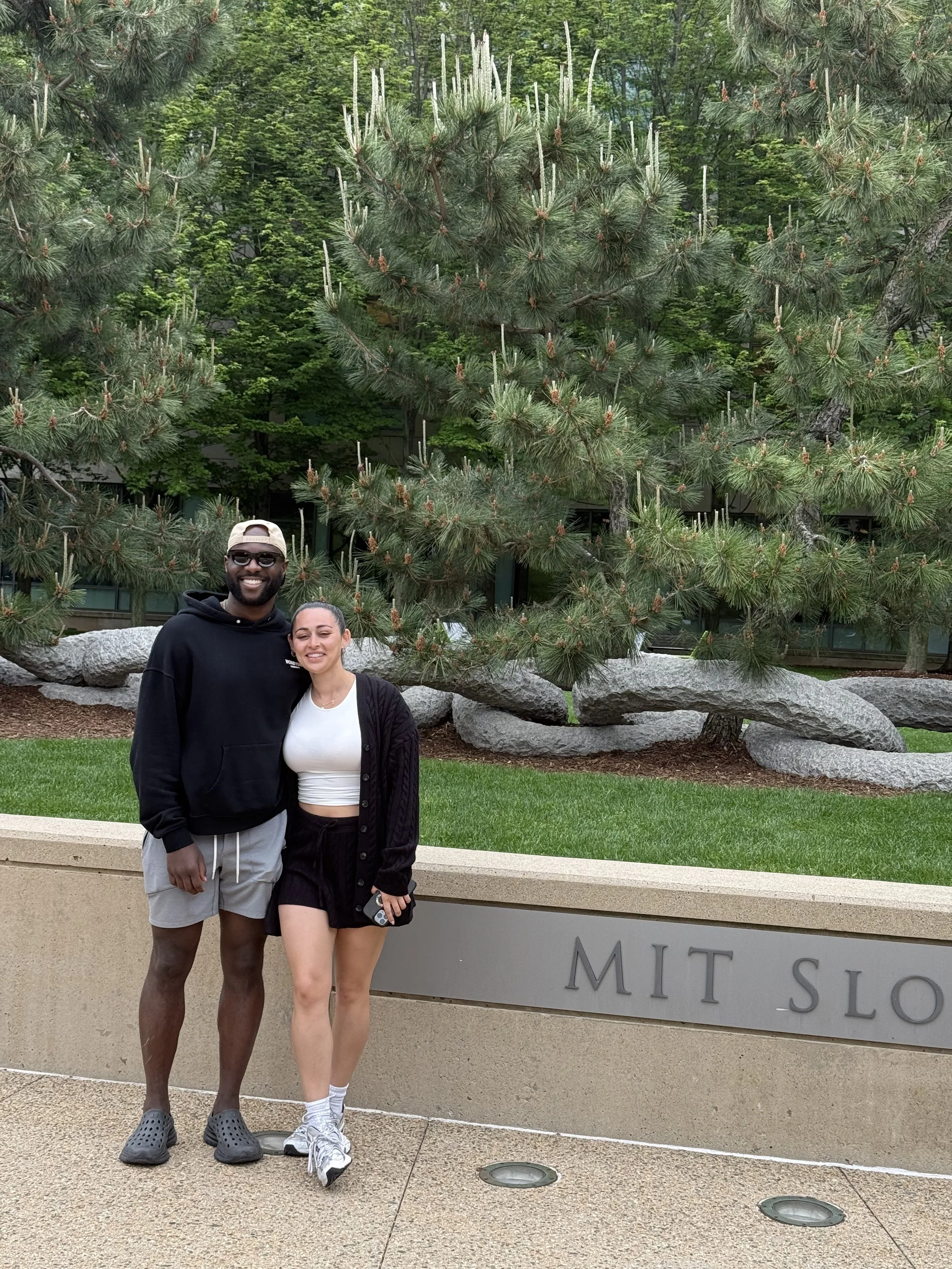 Two people pose in front of a landscaped pine garden and MIT Sloan School sign.