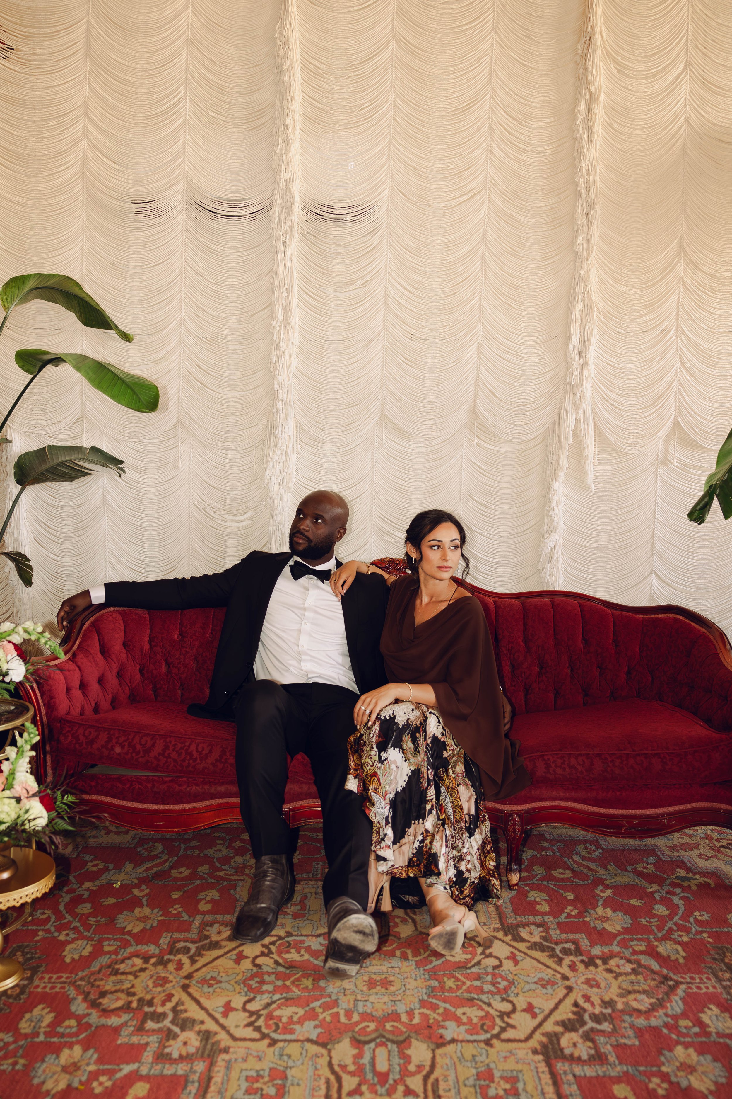 A formally dressed couple sitting on a vintage red velvet sofa in front of a textured cream backdrop with tropical plants.