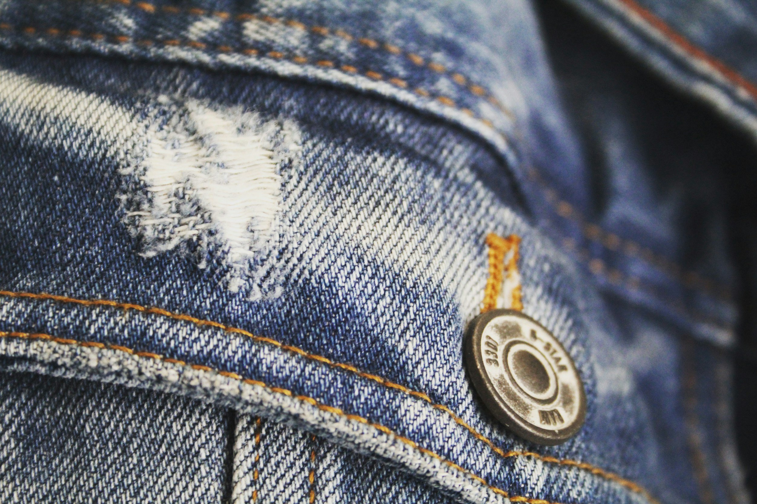 Close-up of blue denim jeans with orange stitching and a metal button, showing a tear and stitching details.