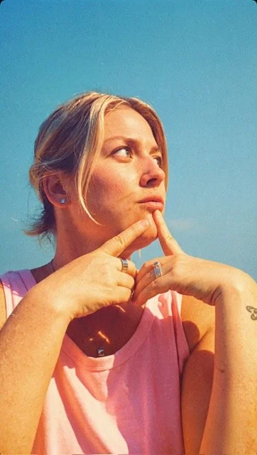 A woman with blonde hair looking to the side, holding her chin with her fingers, against a clear blue sky.