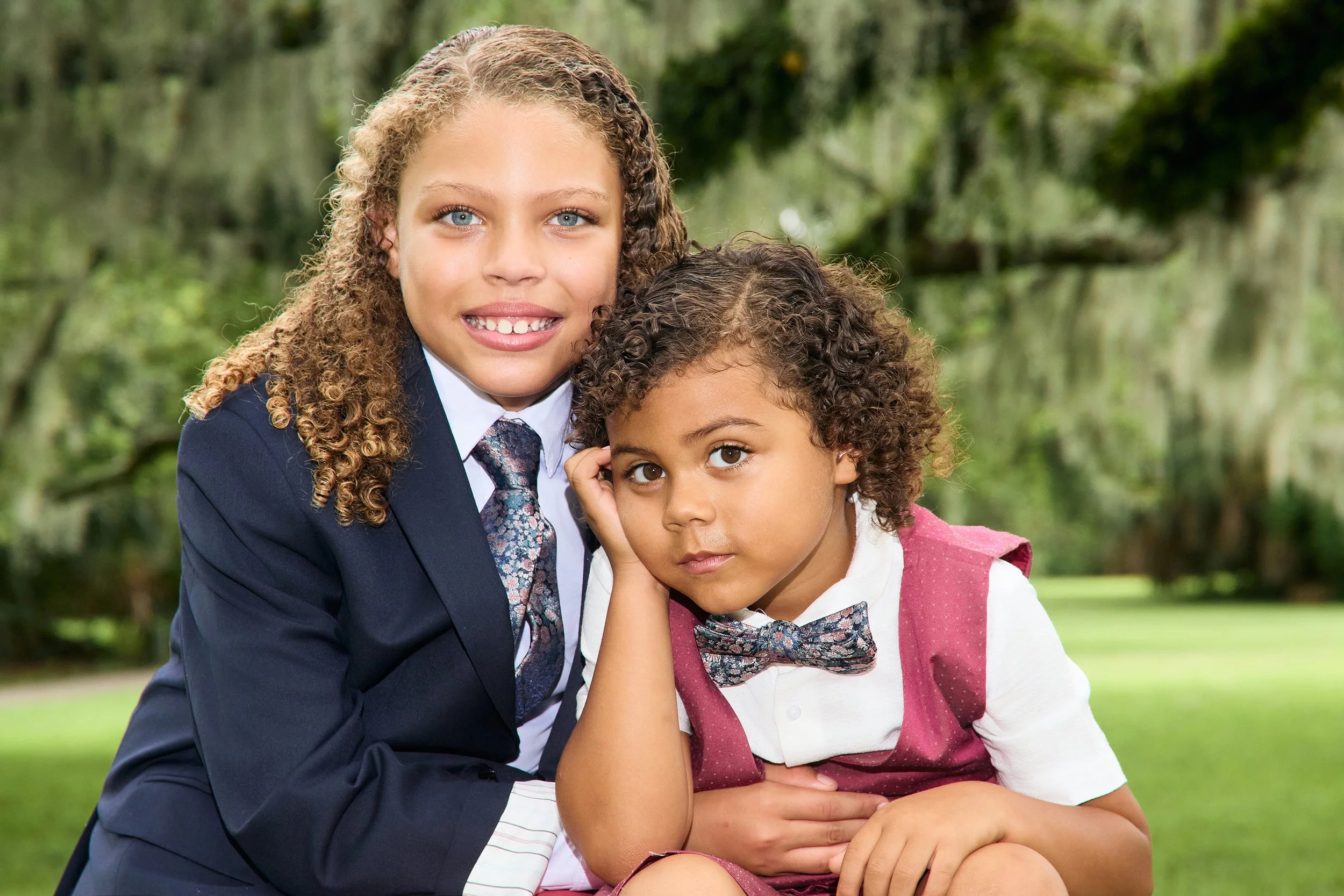 Two children with curly hair in formal attire; one in a suit and tie, the other in a vest and bowtie, sitting outdoors. Shot in New Orleans City Park.