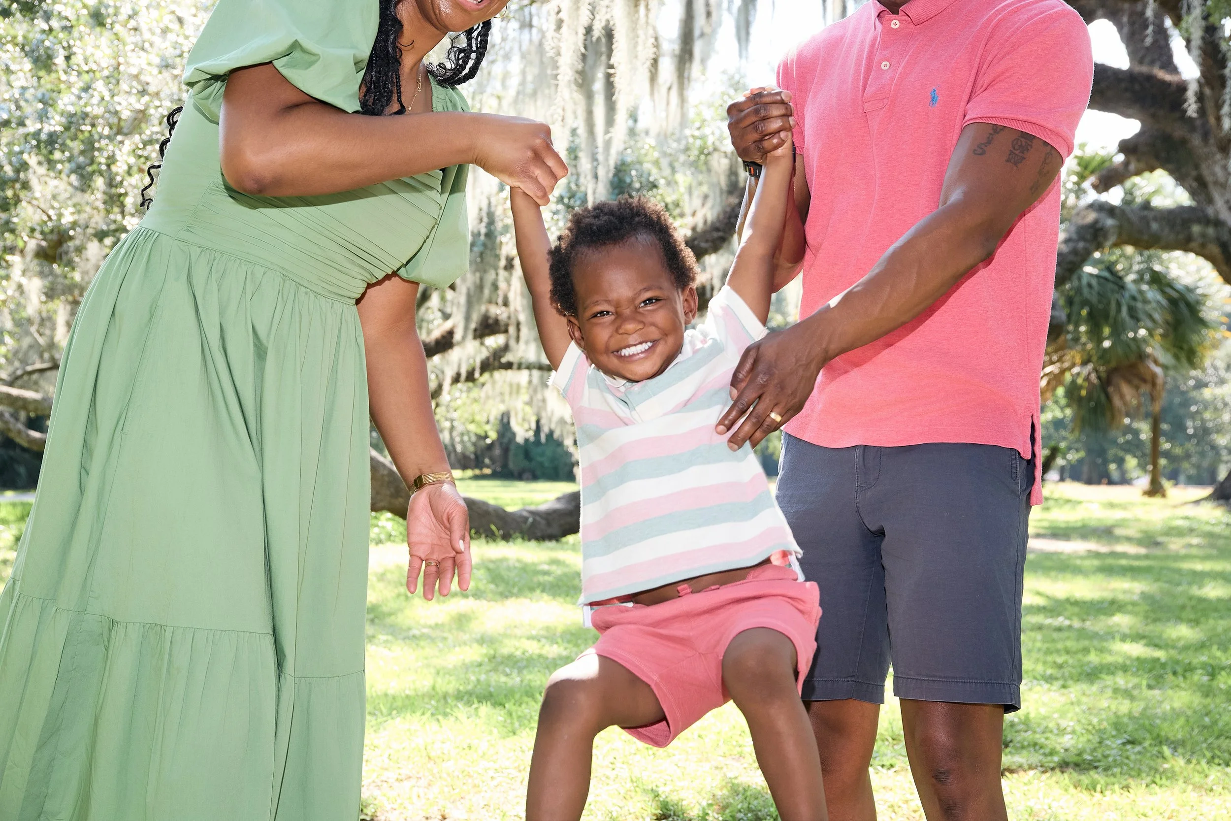 A child smiling while being lifted by two adults in New Orleans City Park with trees in the background.