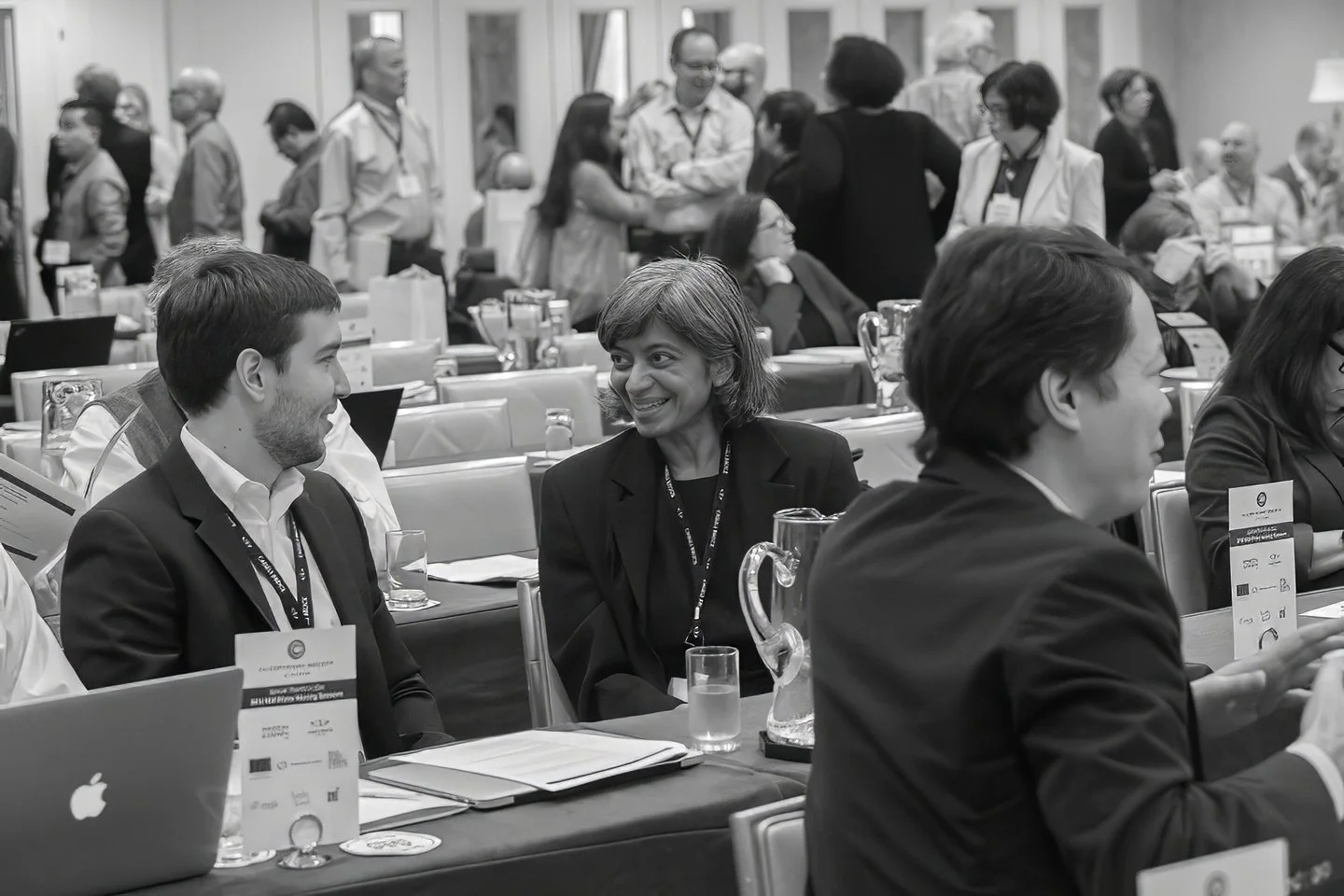 Black and white photo of a conference scene with people sitting at tables, engaged in conversation. There are presentation materials, laptops, and water pitchers on the tables. People in the background are standing and mingling.  Nola conference.