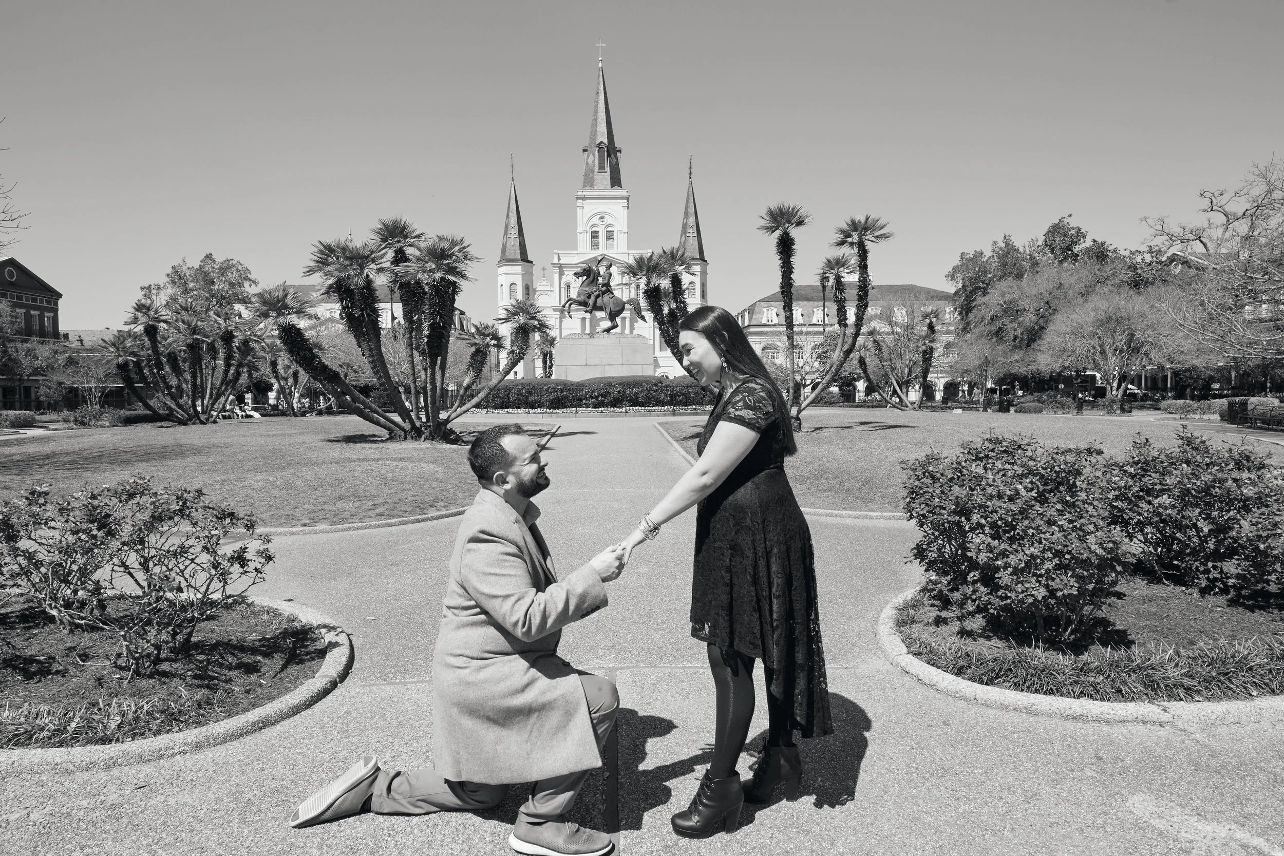 A black and white photo of a man proposing to a woman in Jackson Square. They are in front of St. Louis cathedral with tall spires in the background, surrounded by palm trees and walking paths.
