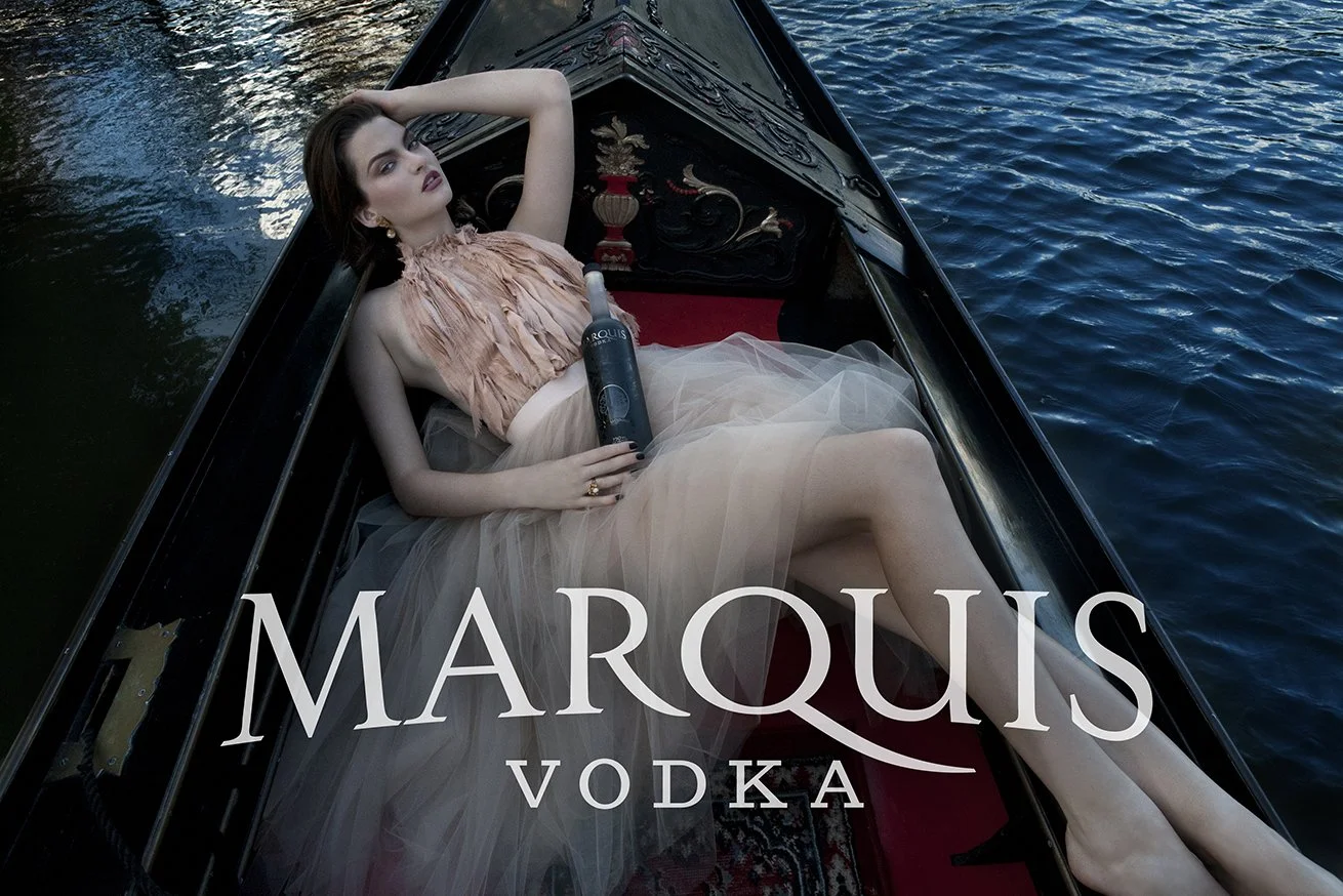 Woman in elegant dress lying in gondola holding Marquis Vodka, fall campaign, with water in background.