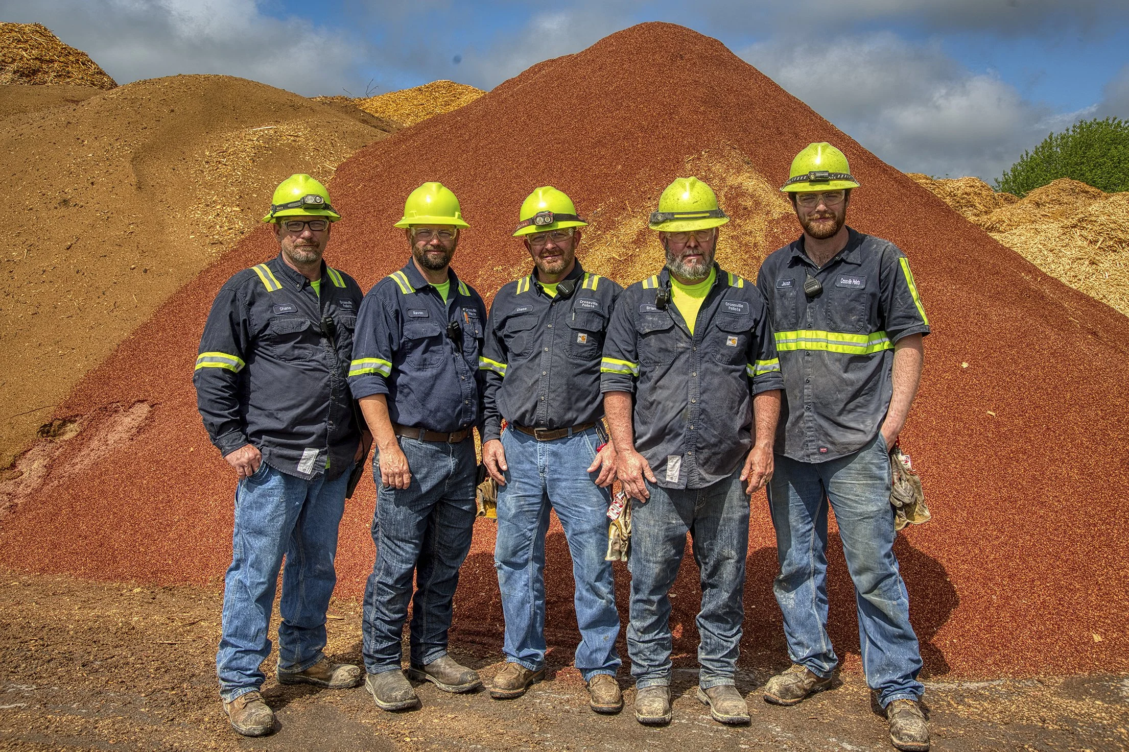 Five industrial workers in safety gear standing in front of piles of mulch.