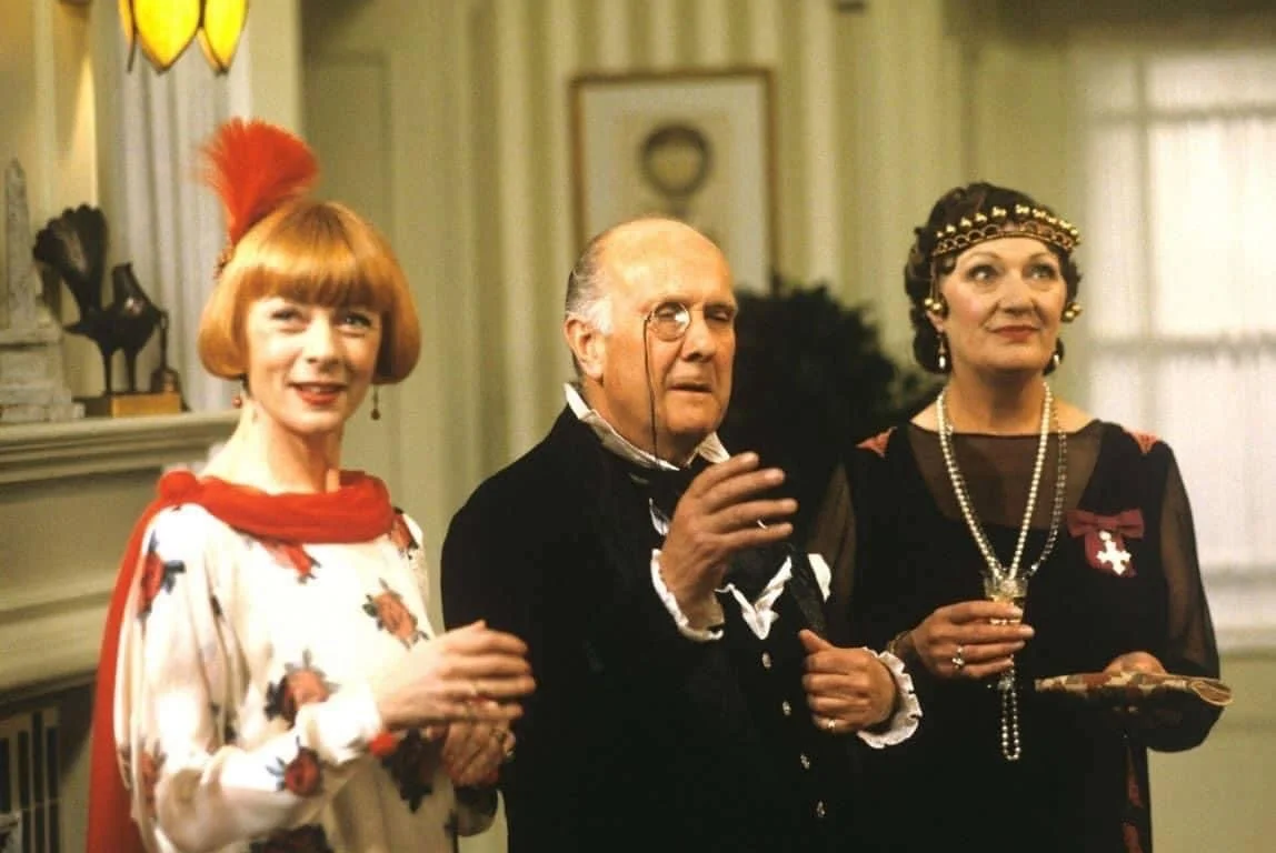 Three people dressed in vintage or costume attire, standing indoors in a living room with a mantel and framed pictures in the background.