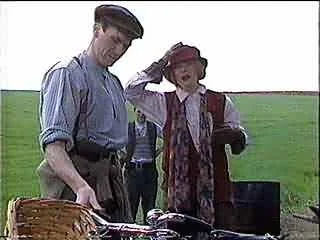 Two women, one in a blue dress and the other in a hat and scarf, standing outdoors next to a table with food and a black box in a grassy field.