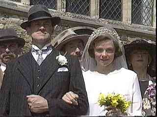 A group of people dressed in historical or formal clothing, with a woman holding flowers, standing outdoors in front of a building.