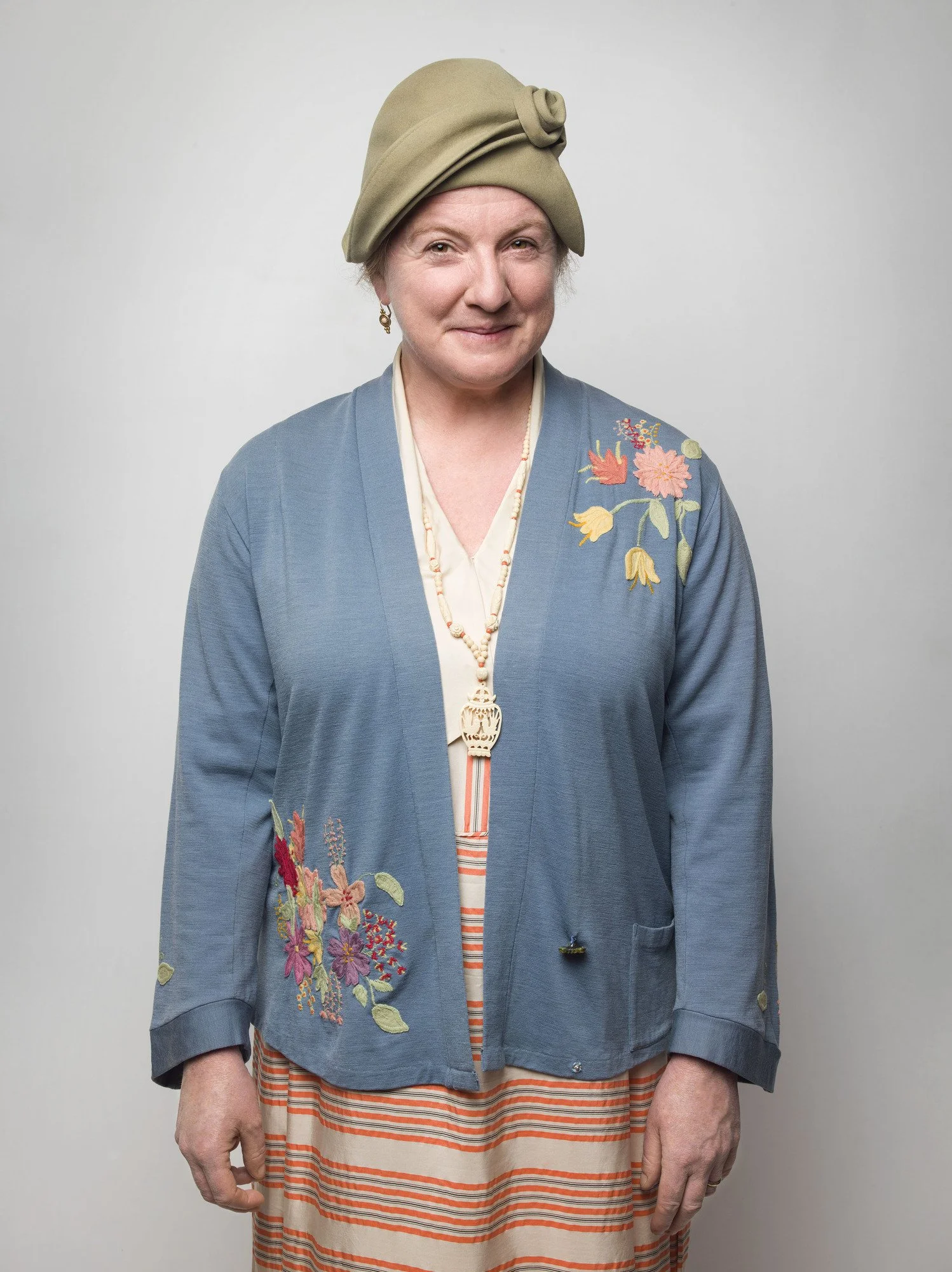 A woman with short gray hair wearing a beige hat with a rose detail, a blue embroidered jacket with floral designs, and a striped skirt, standing against a plain gray background.