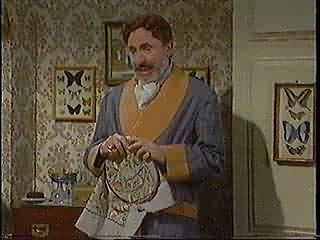 A man with a mustache wearing a gray sweater with yellow trim, holding a paper bag in a room decorated with butterfly artwork.