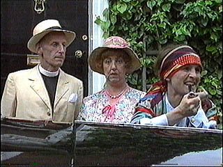 Three women dressed in vintage clothing, with one older woman in a straw hat, another in a floral dress and hat, and the third in a headscarf and striped shirt, sitting in a carriage.