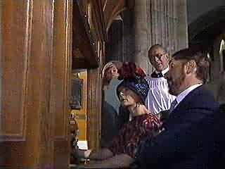 A small group of people gathered around a piano, with some looking at sheet music, in a room with wooden paneling and a stone column.