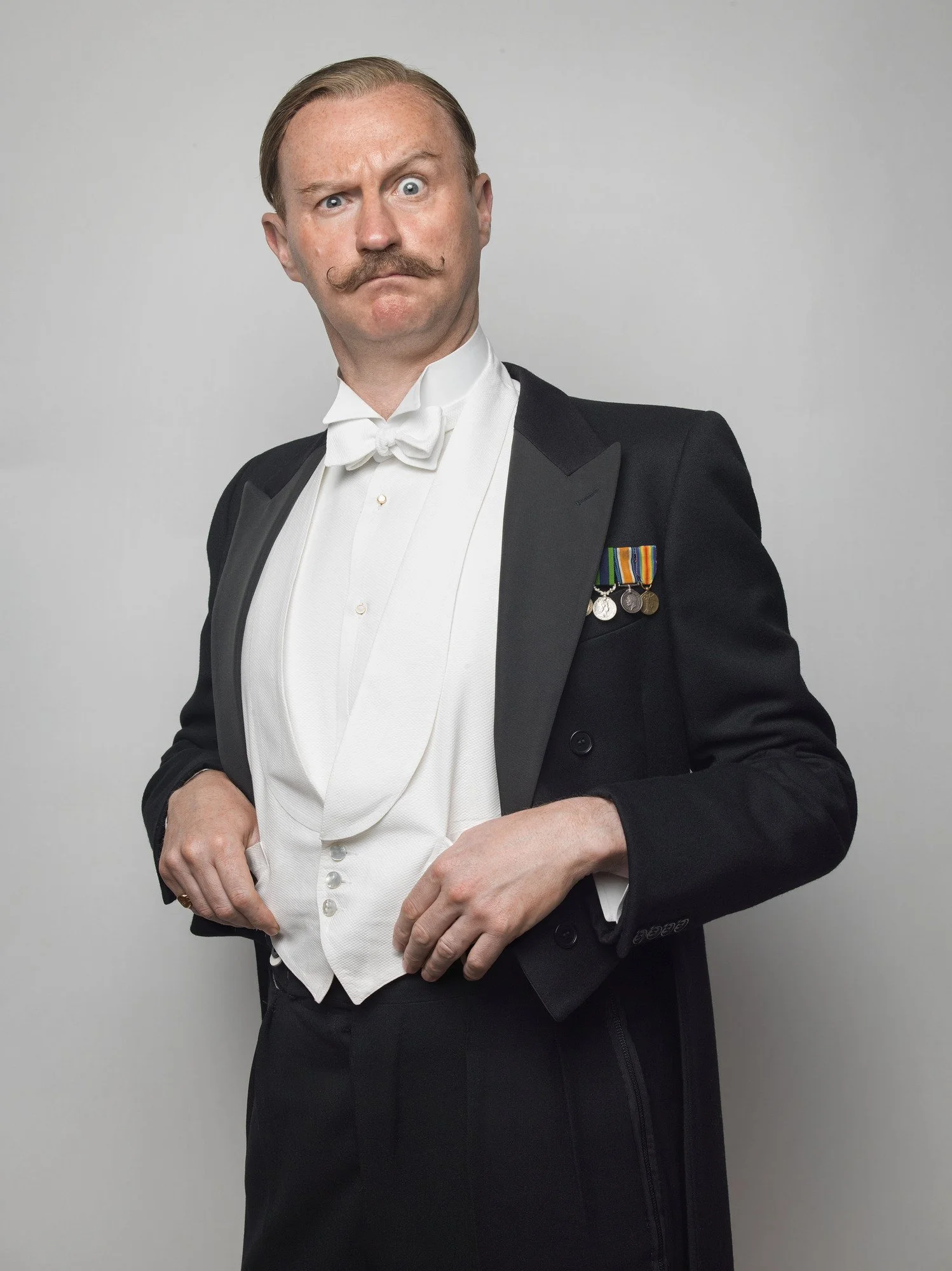 Mark Gatiss dressed in a tuxedo with medals on his chest making a puzzled or skeptical facial expression.