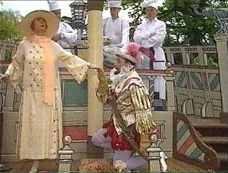 A woman in a yellow dress and hat holds a glove while a clown in colorful costume kneels in front of her. In the background, three people dressed as authorities stand on a porch.