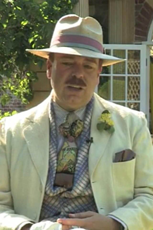 Steve Pemberton in a light-colored suit with a boutonniere and a patterned tie, wearing a safari-style hat, standing outdoors in front of a house with a screened porch and greenery.