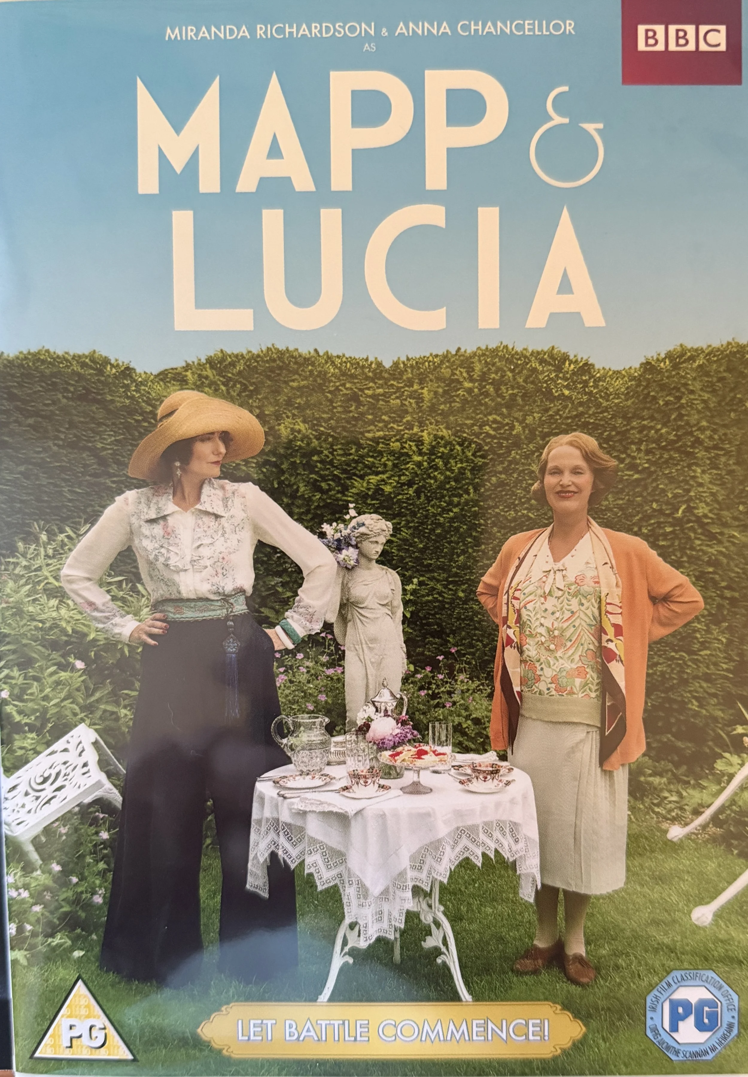 DVD front coverr for a BBC production of 'Mapp & Lucia,' featuring two women dressed in vintage clothing standing outdoors near a garden table with teacups, a teapot, and flowers, with lush green hedges and a clear blue sky in the background.
