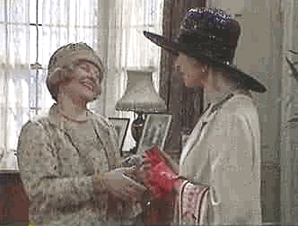 Two women exchanging a red gift in a cozy, vintage-style room with soft lighting, a lamp, and patterned curtains.