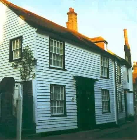 A white two-story house with black window frames and a black front door, with smoke coming out of two brick chimneys. Vines grow along the side of the house.