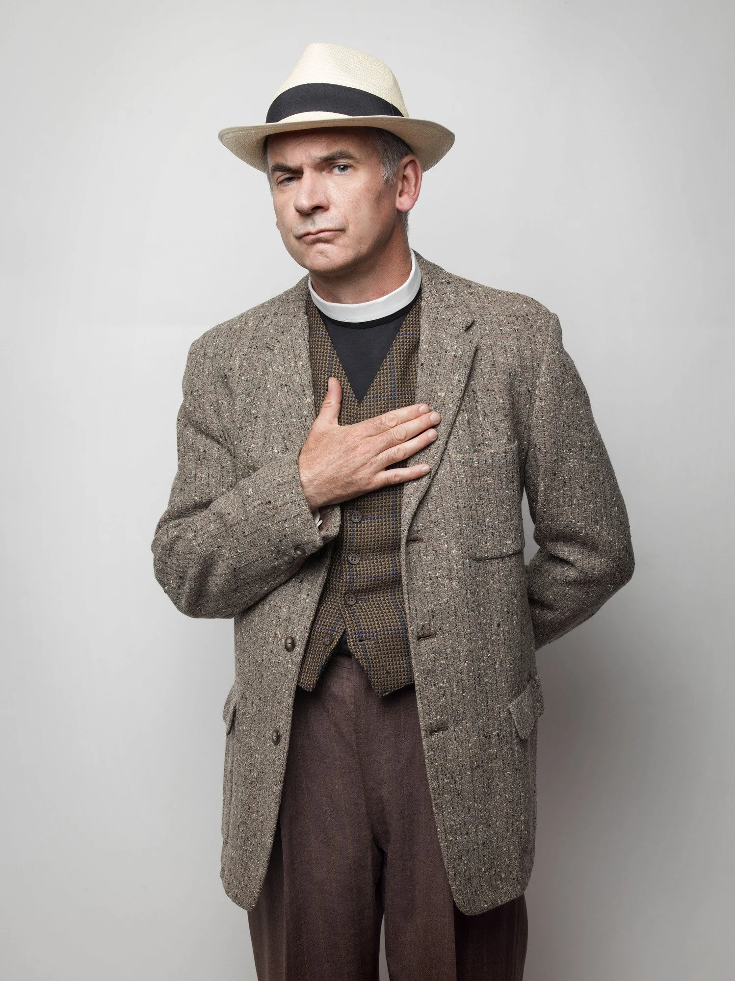 Paul Ritter dressed in vintage style clothing, wearing a tweed overcoat, vest, and brown pants, with a black shirt underneath. He is also wearing a cream-colored fedora.