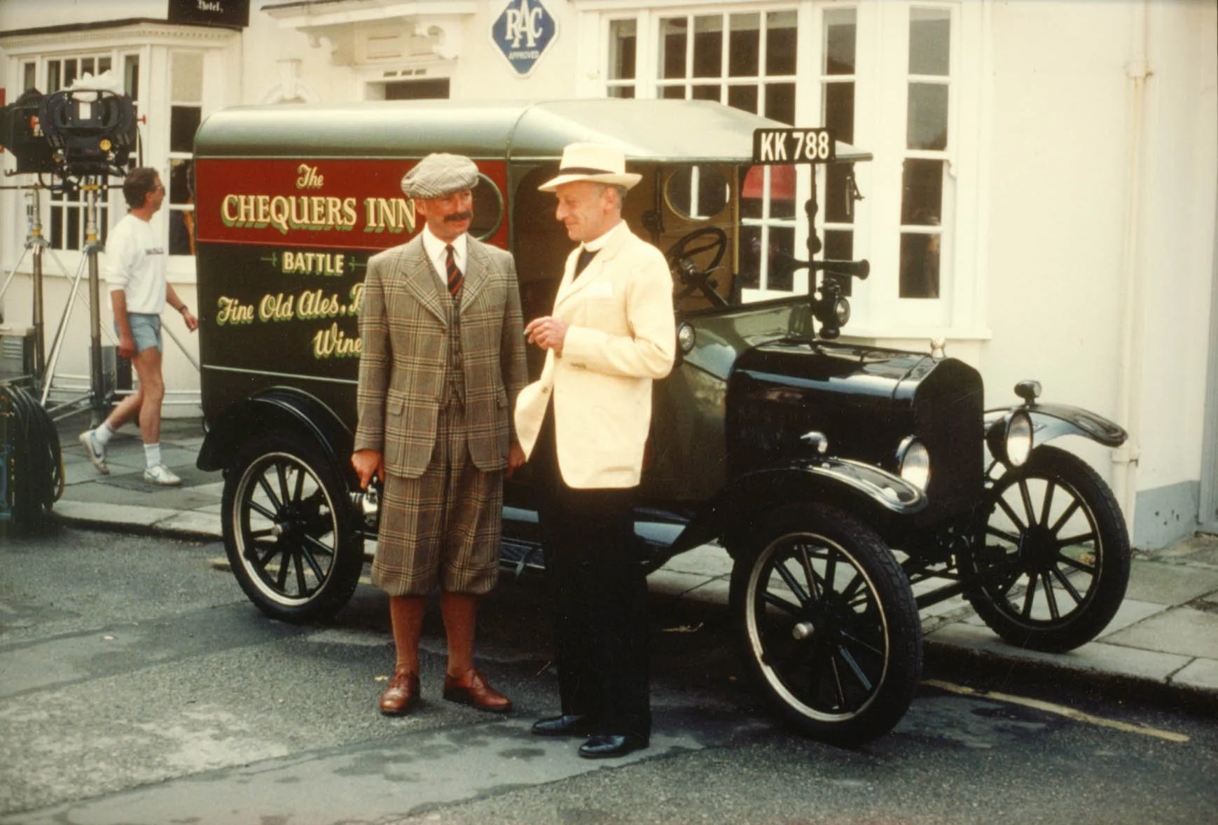 Two men dressed in vintage attire stand in front of a black antique car with a sign that reads "The Chequers Inn" for the TV series Mapp and Lucia