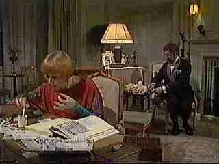 A woman with short blond hair sitting at a cluttered desk in a room with green walls, and a man in a dark suit presenting a tray of food.