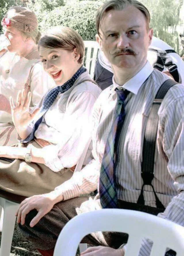 Quaint Irene and Mark Gatiss dressed in vintage or old-fashioned clothing, sitting outdoors. The man in the foreground has a stern expression, and the woman next to him is smiling and waving, while a woman in the background is wearing a hat.