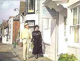 Two people walking down a street past white buildings, one wearing a yellow suit and the other in dark clothing with a hat.