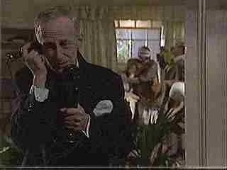 A man in a tuxedo talking on a landline phone indoors with a woman playing guitar in the background.