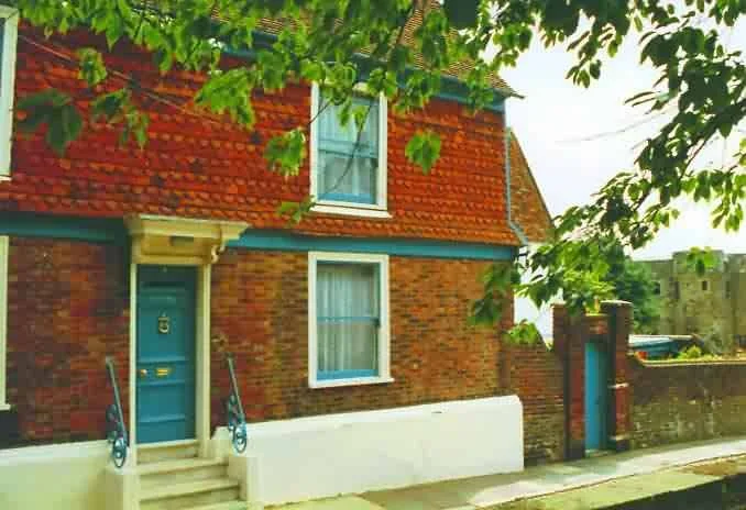 A brick house with a blue front door, white-framed windows, and a tiled roof, partially obscured by green tree branches.