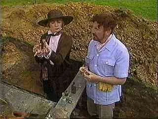 Two women standing at a gravesite, one wearing a black hat and suit, the other in a light blue uniform, holding flowers.