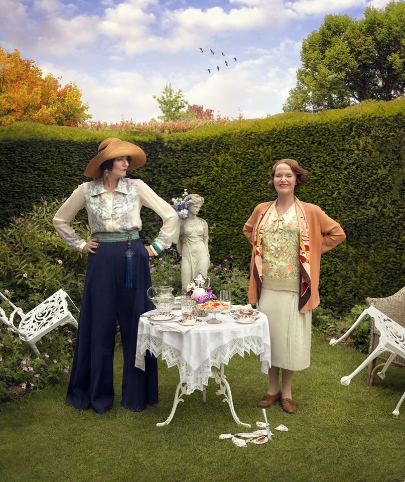 Anna Chancellor and Miranda Richardson stand outdoors in a garden among lush greenery and colorful trees, with a table set for tea and a statue between them.