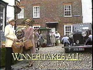 People gathering on a cobblestone street near a vintage car, with a brick building in the background, and a sign that reads 'WINNER TAKES ALL'.