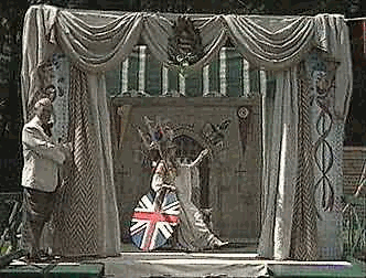 A performer in a white and blue costume with a Union Jack design is on stage, holding a microphone, with a man in a suit standing nearby. The stage is decorated with curtains and a backdrop.