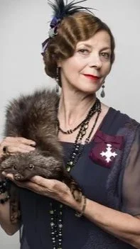 An older woman dressed in vintage clothing holding a small furry animal, possibly a ferret or similar creature, against a plain background.