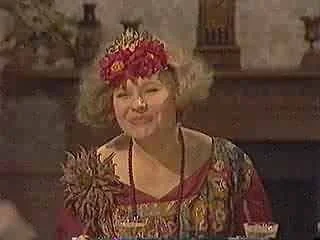 Older woman with curly blonde hair wearing a floral dress and a red floral headband, smiling in a cozy room.