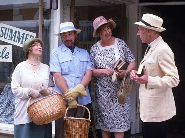 Four people dressed in vintage clothing, three women and one man, are standing outside a store having a conversation. The women are holding baskets and books, and all are wearing hats.