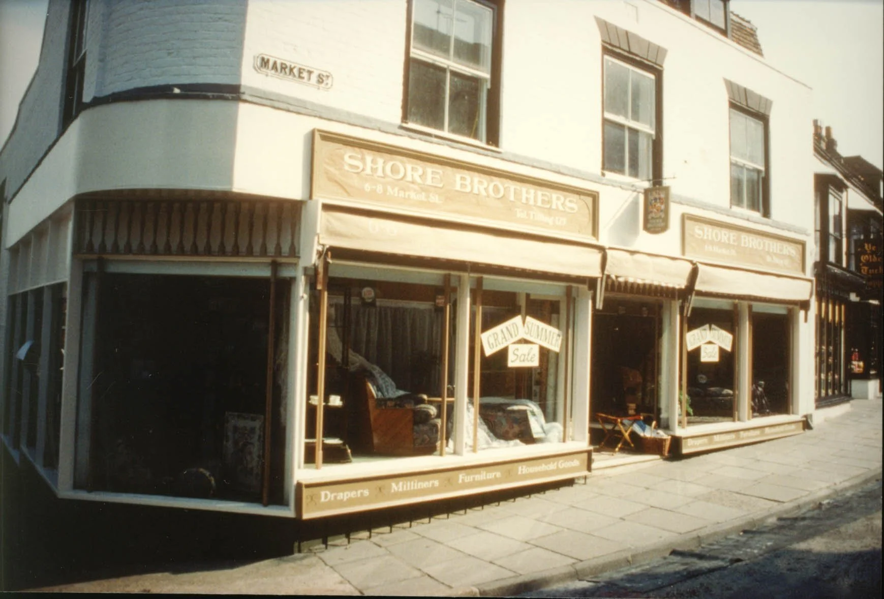 Corner view of Shore Brothers storefront on Market Street, with large display windows and a sign advertising a grand summer sale, located at 6-8 Market Street, with residential windows above and neighboring shops on either side.