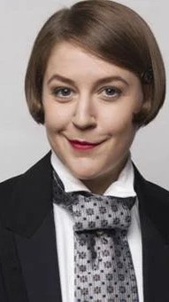 A woman with short brown hair, wearing a black blazer, white shirt, and a patterned bow tie, smiling slightly against a gray background.