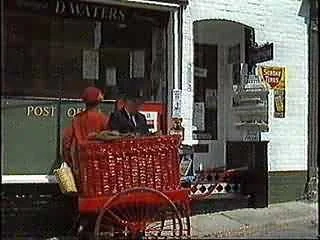 Person dressed as Santa Claus sitting at a table outside a store, with a person in a red hat standing nearby.