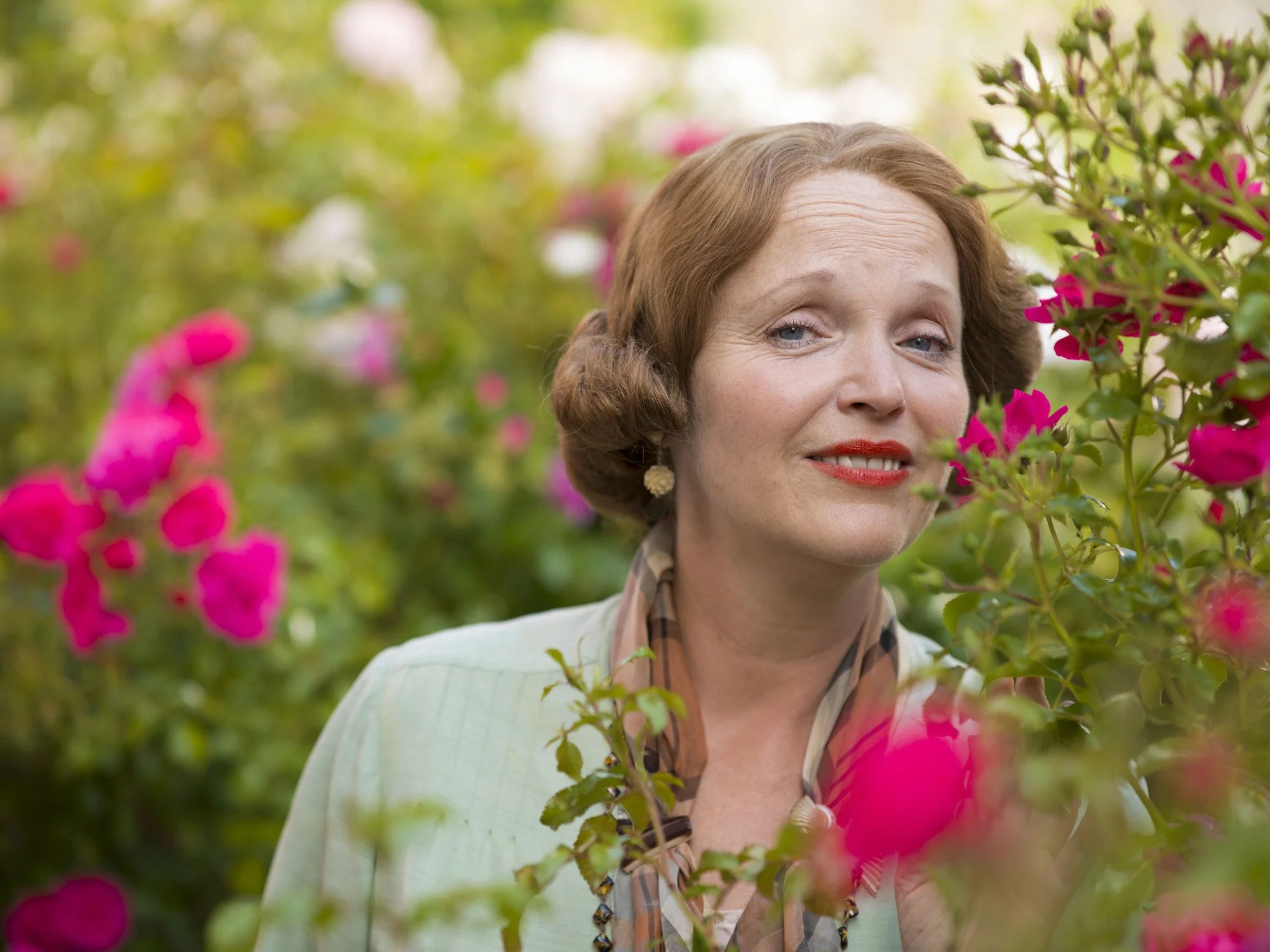 Miranda Richardson  smiling among pink flowers in a garden setting.