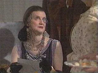 A woman with dark hair, glasses, and wearing a fancy dress with jewelry sitting at a table.