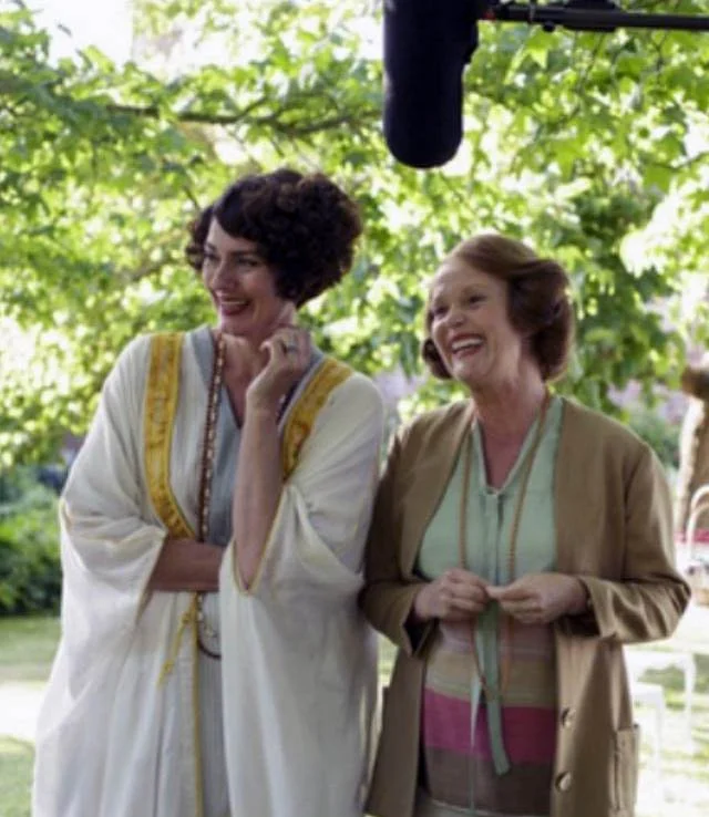 Mapp and Lucia  smiling and laughing outdoors under green tree leaves, one wearing a white dress with yellow embroidery, the other in a beige jacket with a striped dress underneath.